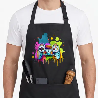 Tablier de cuisine noir avec une manette de jeu vidéo multicolore et des éclaboussures de peinture stylisées.