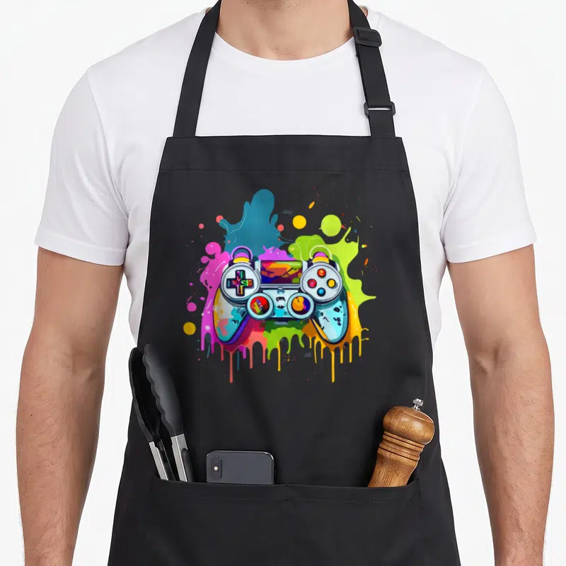 Tablier de cuisine noir avec une manette de jeu vidéo multicolore et des éclaboussures de peinture stylisées.