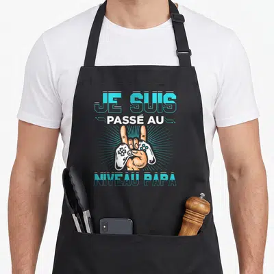 Tablier de cuisine noir avec inscription Je suis passé au niveau papa et main tenant une manette blanche.