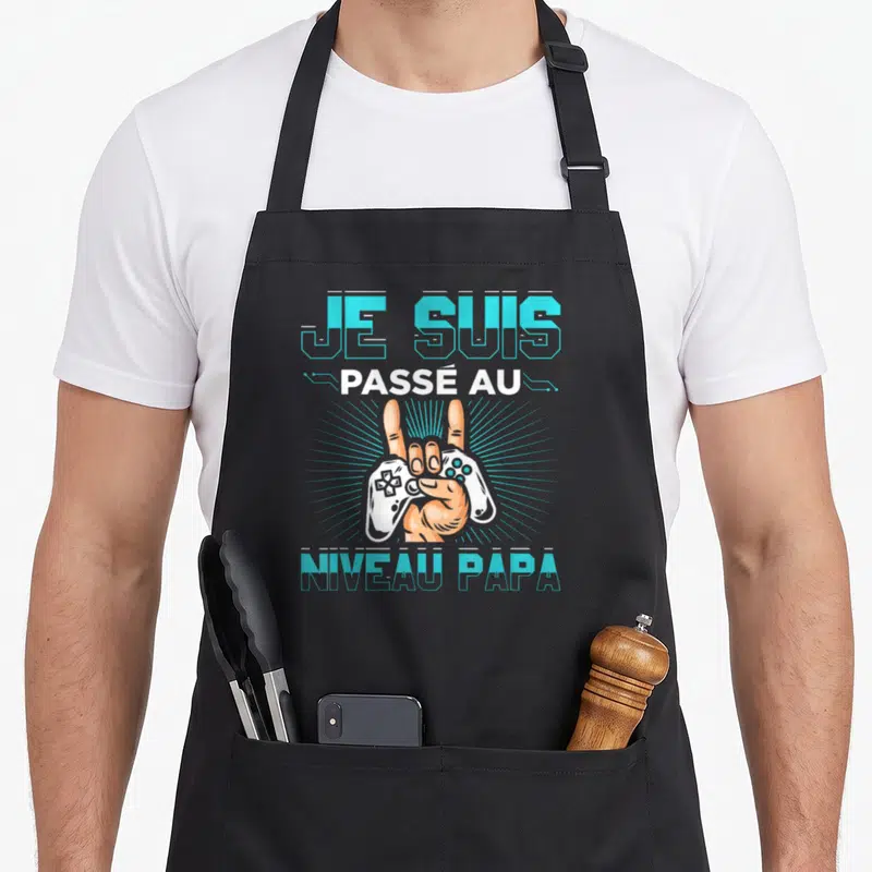 Tablier de cuisine noir avec inscription Je suis passé au niveau papa et main tenant une manette blanche.