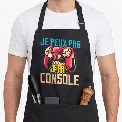 Tablier de cuisine noir avec une manette rouge et le texte Je peux pas j'ai console imprimé en couleur.