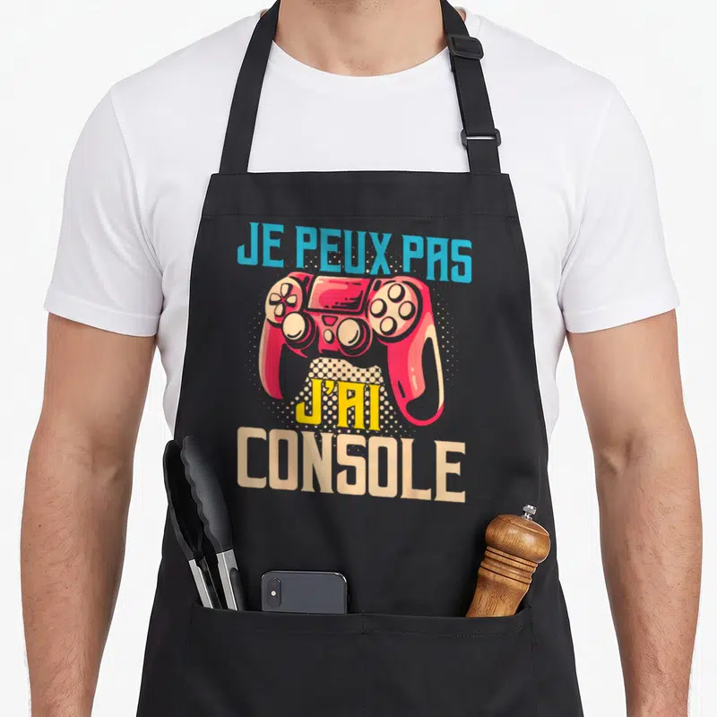 Tablier de cuisine noir avec une manette rouge et le texte Je peux pas j'ai console imprimé en couleur.