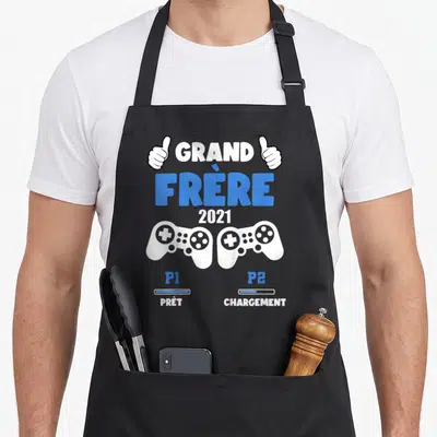 Tablier de cuisine noir avec texte Grand Frère 2021 et deux manettes de console blanches.