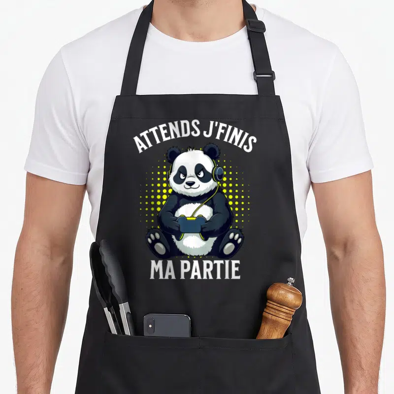Tablier de cuisine noir avec illustration d'un panda gamer portant des écouteurs et tenant une manette de jeu vidéo sur fond à pois jaunes.