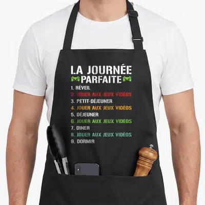 Tablier de cuisine noir avec une liste humoristique intitulée La journée parfaite et des icônes de manettes vertes.