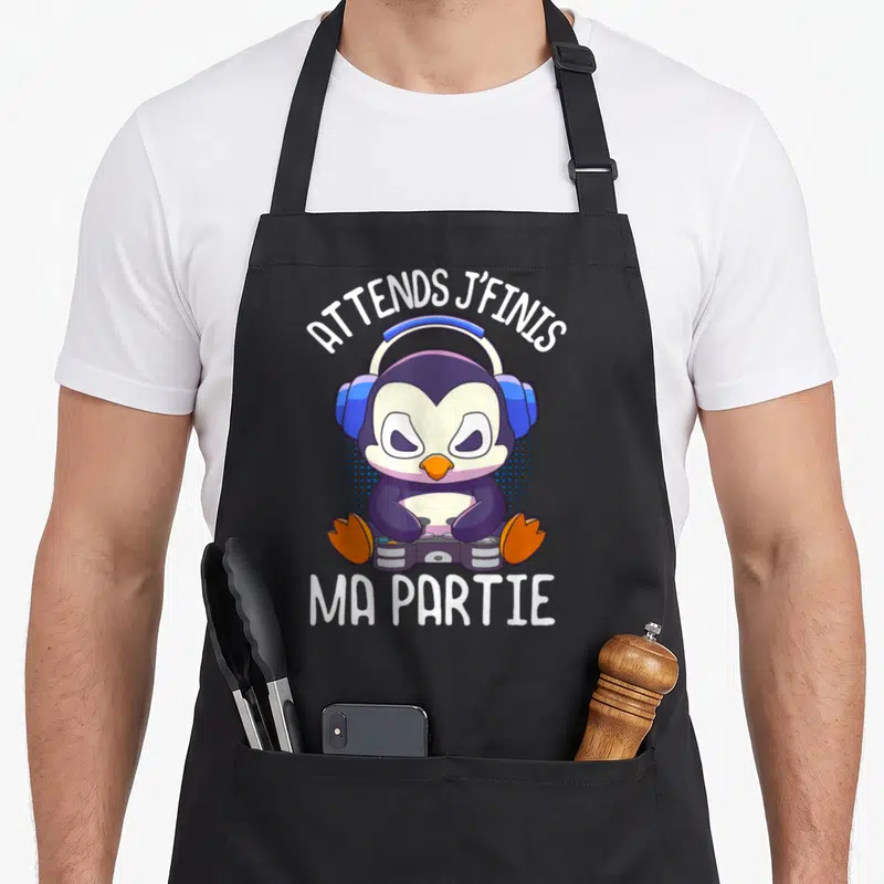 Tablier de cuisine noir avec manchot gamer aux écouteurs et texte blanc Attends j'finis ma partie