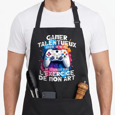 Tablier de cuisine noir avec manette de jeu vidéo et texte humoristique sur fond de peinture éclaboussée.