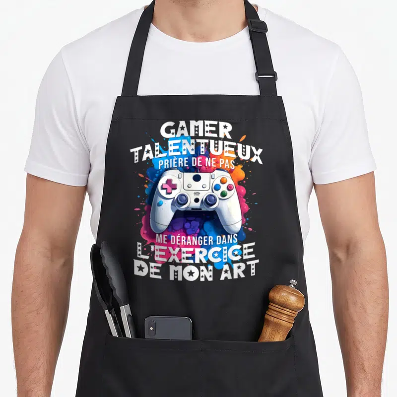 Tablier de cuisine noir avec manette de jeu vidéo et texte humoristique sur fond de peinture éclaboussée.
