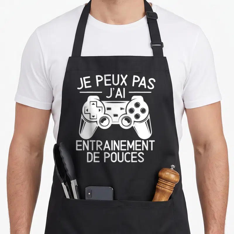 Tablier de cuisine noir avec une illustration de manette de jeu et le texte Je peux pas j'ai entrainement de pouces en blanc.