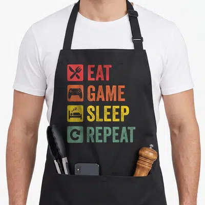 Tablier de cuisine noir avec les mots Eat Game Sleep Repeat et des icônes colorées.