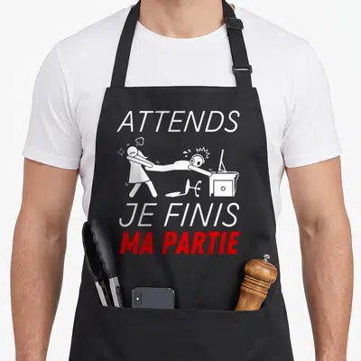 Tablier de cuisine noir avec texte blanc et rouge Attends Je Finis Ma Partie et dessin d'une dispute familiale.