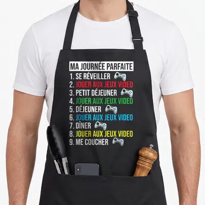 Tablier de cuisine noir avec liste humoristique Ma Journée Parfaite et illustrations de manettes de jeu colorées.