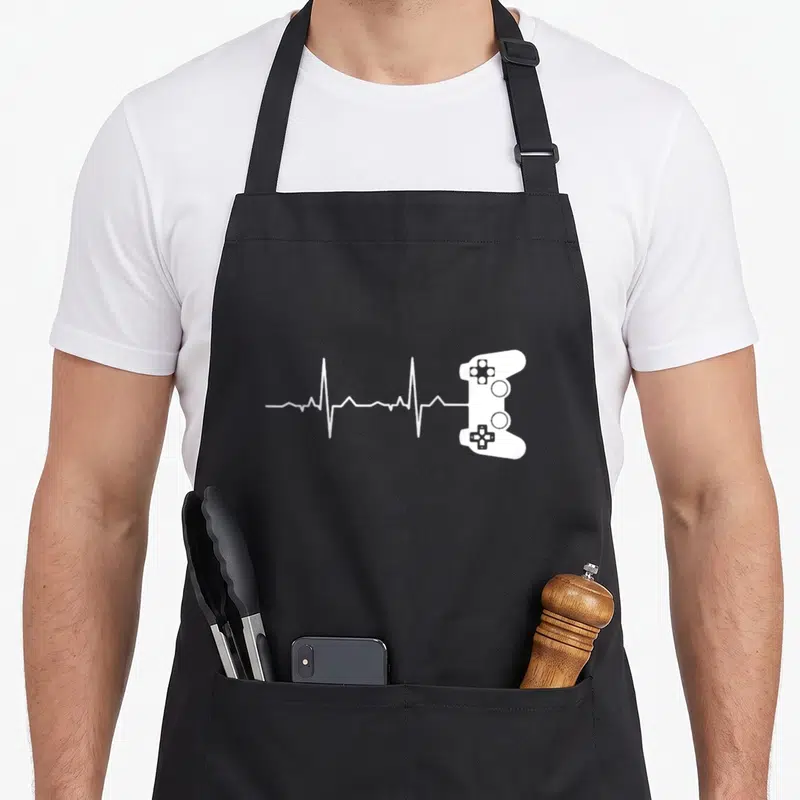 Tablier de cuisine noir avec une ligne de vie blanche se terminant par une manette de jeu vidéo.