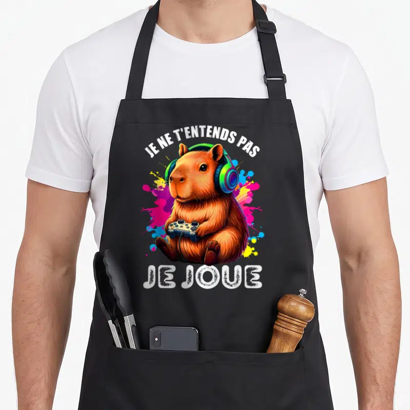 Tablier de cuisine noir avec un capybara tenant une manette et portant des écouteurs colorés.