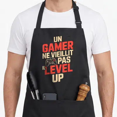 Tablier de cuisine noir avec le texte Un gamer ne vieillit pas il level up et une illustration de manette de jeu.