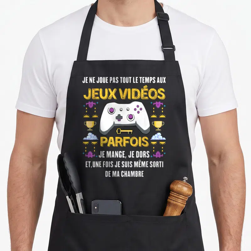 Tablier de cuisine noir avec manette blanche, texte jaune et motifs pixel art rétro sur fond uni.