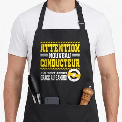 Tablier de cuisine noir avec le texte Attention Nouveau Conducteur J'ai tout appris grace au gaming et une manette jaune.