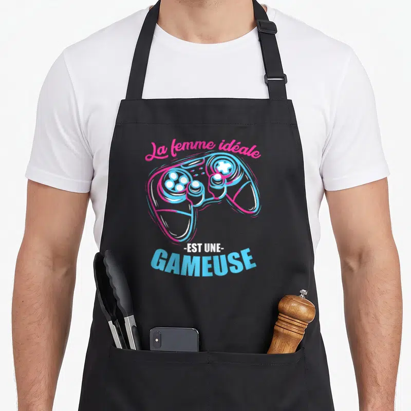 Tablier de cuisine noir avec illustration colorée d'une manette de jeu et texte humoristique en rose et bleu.