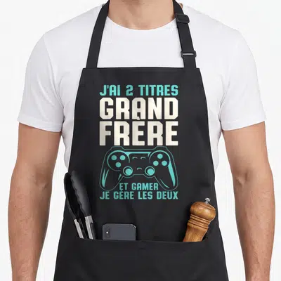 Tablier de cuisine noir avec inscription J'ai 2 titres Grand Frère et illustration de manette de jeu vidéo turquoise.