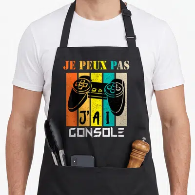 Tablier de cuisine noir avec manette de jeu et texte Je peux pas j'ai console sur fond de bandes colorées.