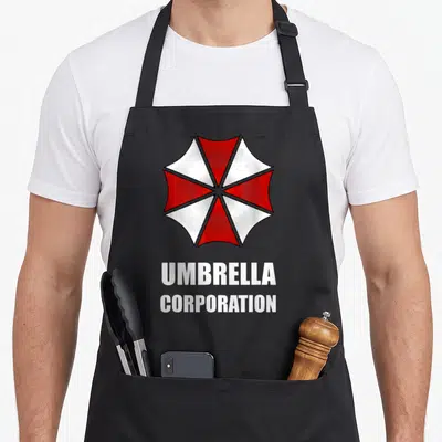 Tablier de cuisine noir avec logo parapluie rouge blanc Umbrella Corporation et texte blanc en dessous.
