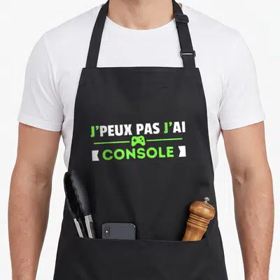 Tablier de cuisine noir avec le texte J'peux pas j'ai console et une icône de manette vert lime au centre.