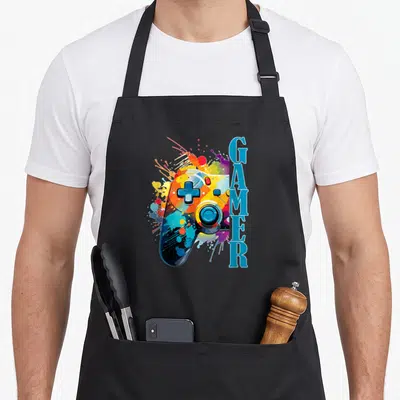 Tablier de cuisine noir avec une illustration de manette de jeu vidéo aux couleurs vives et le texte GAMER en bleu sur le côté.