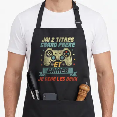 Tablier de cuisine noir avec manette de jeu et texte J'ai 2 titres grand frère et gamer sur fond uni.