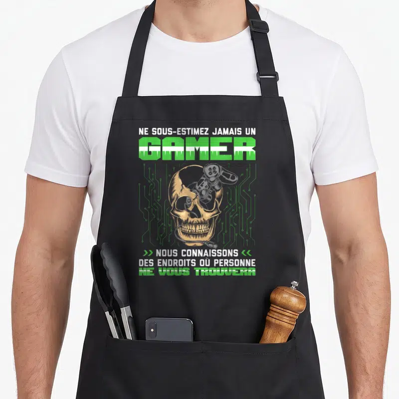Tablier de cuisine noir orné d'un crâne doré sur fond de circuits verts et du texte Ne sous-estimez jamais un gamer.