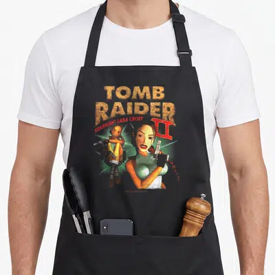 Tablier de cuisine noir avec illustration vintage de Lara Croft tenant des pistolets et le texte Tomb Raider II Starring en lettres texturées.