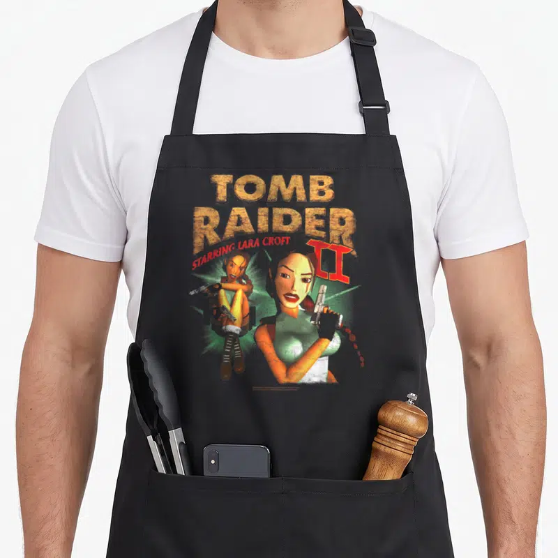 Tablier de cuisine noir avec illustration vintage de Lara Croft tenant des pistolets et le texte Tomb Raider II Starring en lettres texturées.