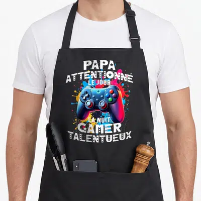 Tablier de cuisine noir avec illustration de manette de jeu et texte Papa gamer talentueux sur fond de taches colorées.