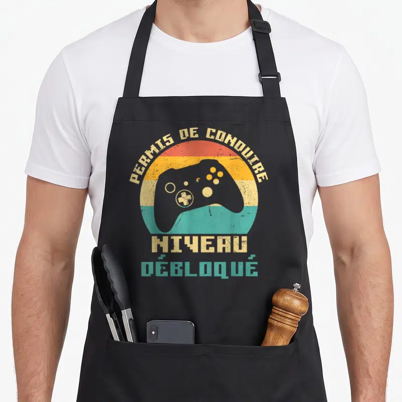 Tablier de cuisine noir avec une manette de jeu vidéo et le texte Permis de Conduire Niveau Débloqué sur fond coloré vintage.