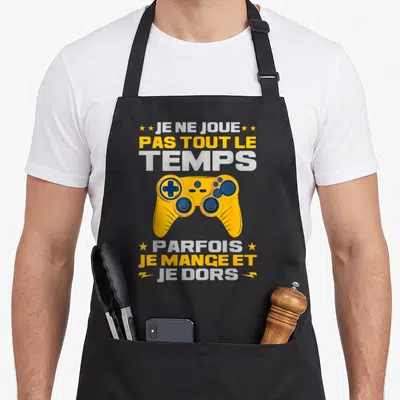 Tablier de cuisine noir avec manette jaune et texte Je ne joue pas tout le temps parfois je mange et je dors.