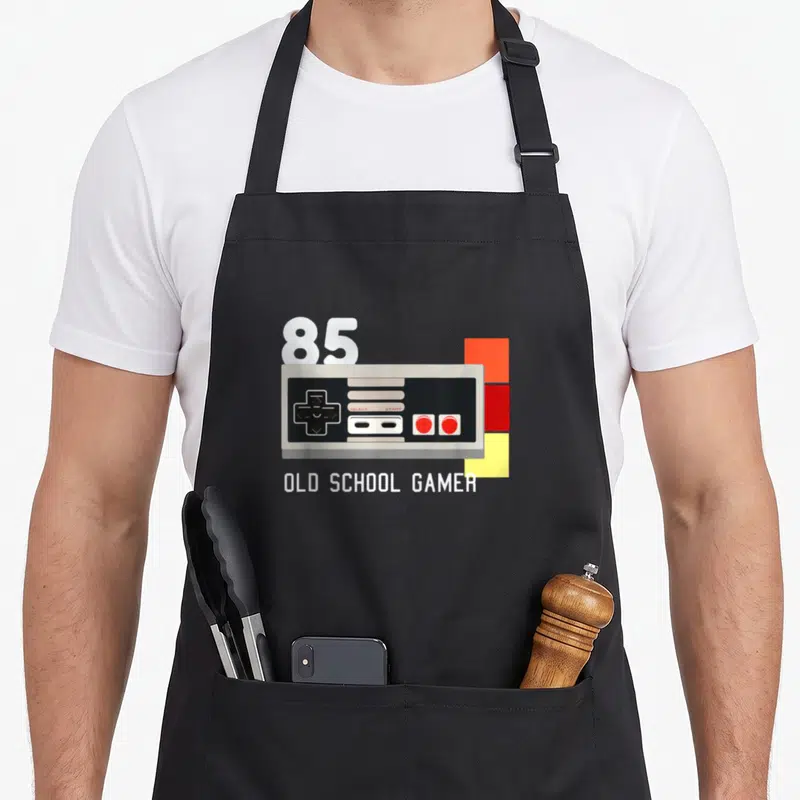 Tablier de cuisine noir avec illustration de manette rétro, le chiffre 85 et Old School Gamer en blanc.