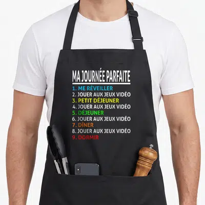 Tablier de cuisine noir avec texte Ma Journée Parfaite listant jouer aux jeux vidéo à plusieurs reprises en couleurs vives.
