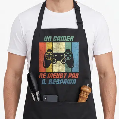 Tablier de cuisine noir avec manette de jeu colorée et texte Un Gamer Ne Meurt Pas Il Respawn imprimé en style vintage.