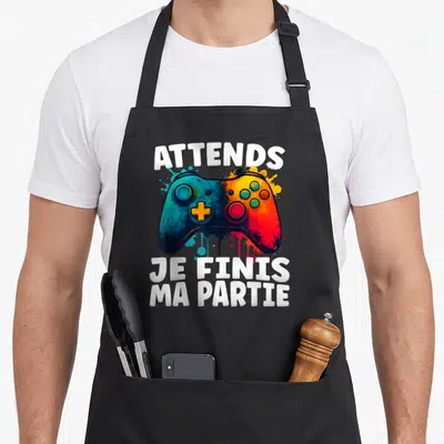 Tablier de cuisine noir avec une manette de jeu multicolore et le texte Attends Je Finis Ma Partie en blanc.