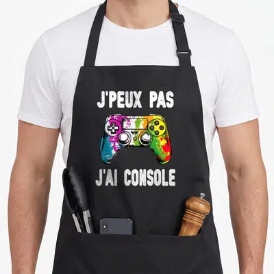 Tablier de cuisine noir avec une manette de jeu vidéo multicolore et le texte J'peux pas j'ai console imprimé en blanc.