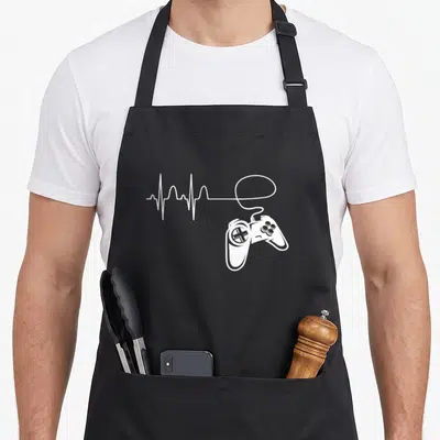 Tablier de cuisine noir avec une illustration blanche représentant une manette de jeu et un tracé de rythme cardiaque stylisé en électrocardiogramme.