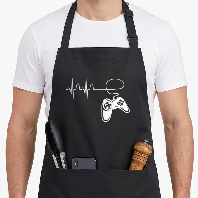 Tablier de cuisine noir avec une illustration blanche représentant une manette de jeu et un tracé de rythme cardiaque stylisé en électrocardiogramme.