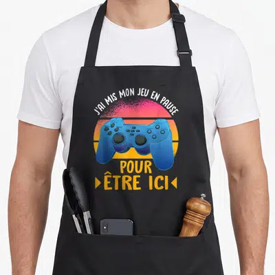 Tablier de cuisine noir avec manette de console bleue et texte blanc sur fond de soleil rétro orange et rose.