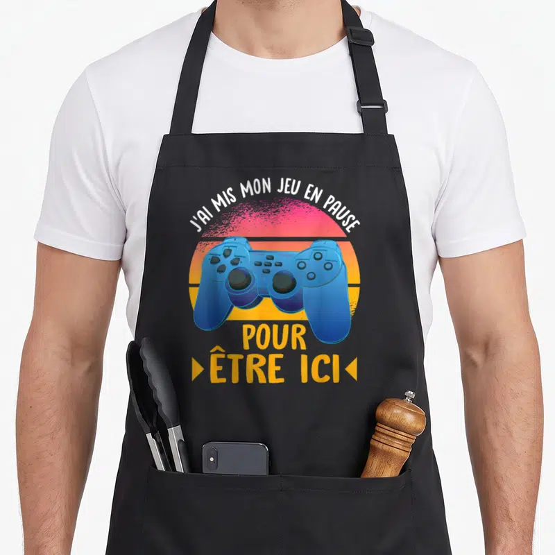 Tablier de cuisine noir avec manette de console bleue et texte blanc sur fond de soleil rétro orange et rose.