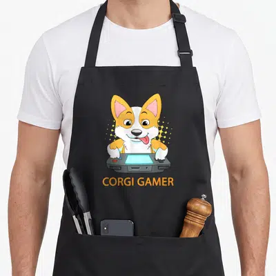 Tablier de cuisine noir avec illustration colorée d'un corgi tenant une console de jeu vidéo et le texte Corgi Gamer en bas.