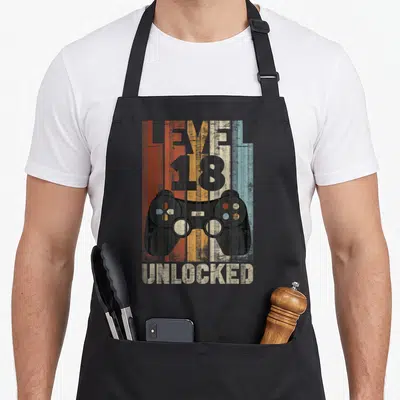 Tablier de cuisine noir avec illustration manette rétro et texte Niveau 18 Débloqué Unlocked sur fond texturé multicolore.