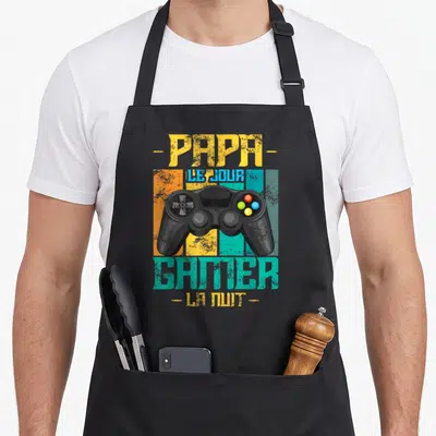 Tablier de cuisine noir avec illustration de manette de jeu et texte Papa Le Jour Gamer La Nuit en couleurs vives.