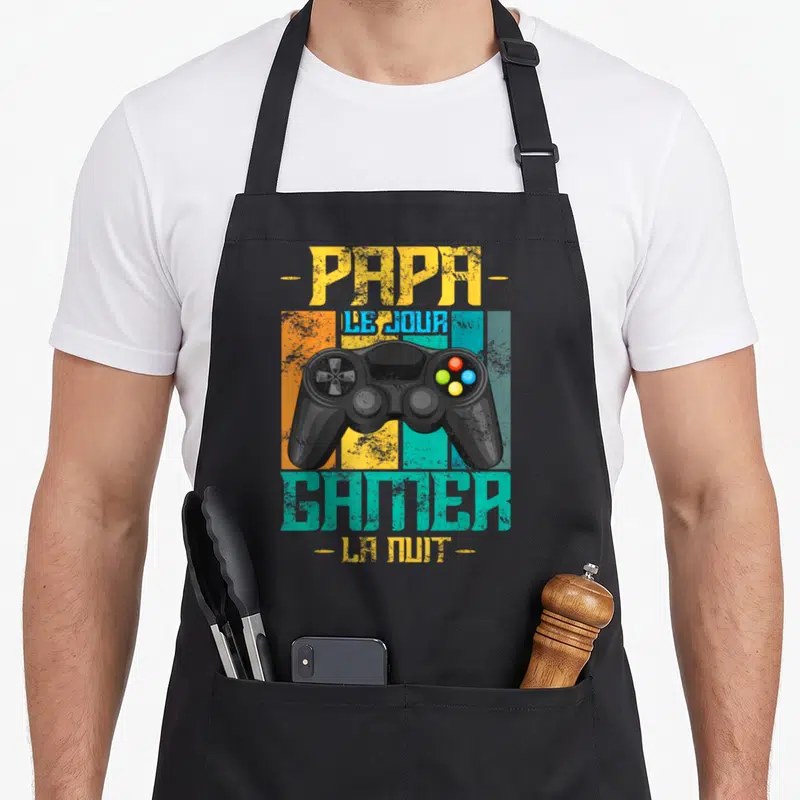 Tablier de cuisine noir avec illustration de manette de jeu et texte Papa Le Jour Gamer La Nuit en couleurs vives.