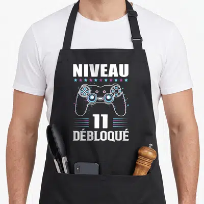 Tablier de cuisine noir avec une manette de jeu vidéo blanche et l'inscription Niveau 11 Débloqué en lettres capitales.