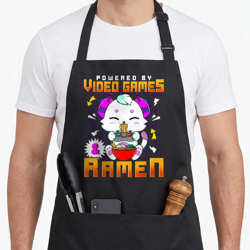 Tablier de cuisine noir avec un chat blanc gamertenant une manette et un bol de ramen rouge sur fond quadrillé violet.