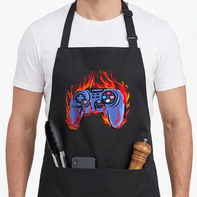 Tablier de cuisine noir avec une illustration de manette de jeu vidéo bleue consumée par des flammes rouges et jaunes vives.